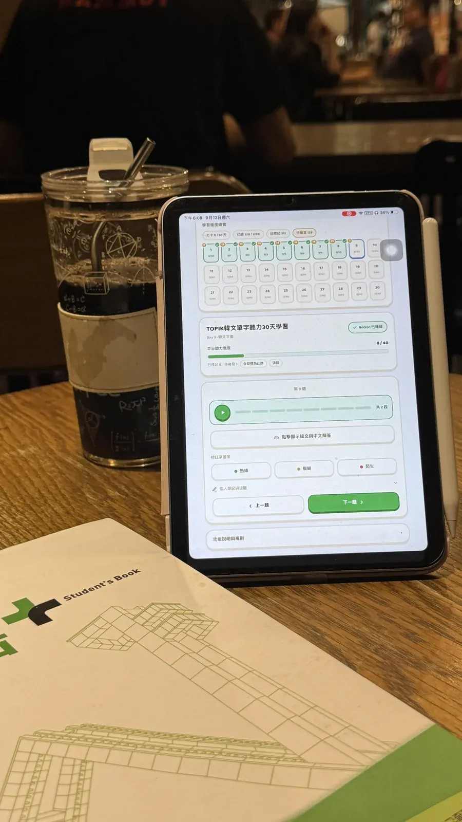 咖啡廳桌上立著 iPad 顯示 TOPIK 單字盲聽打卡工具 旁邊是飲料杯與韓文課本